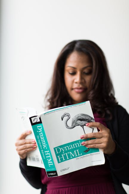 A woman with long dark hair, dressed in a black blazer and maroon top, is holding an open book titled 'Dynamic HTML' by O'Reilly, which features a detailed illustration of a flamingo on the cover. She