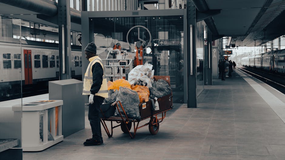 A man wearing a dark beanie, black jacket, and a high-visibility vest stands on a train station platform beside a large, red, metal utility cart loaded with various bags and bundled rubbish, including